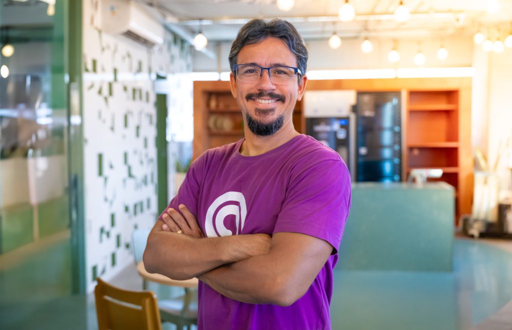 Retrato de homem de braços cruzados sorrindo em escritório moderno, com camisa roxa da Agilize.