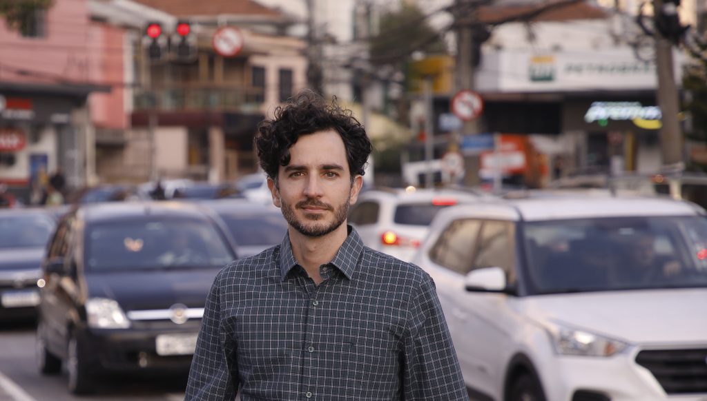 Retrato de homem em pé em avenida movimentada, com carros ao fundo em horário de pico.