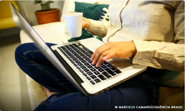 Pessoa sentada no sofá usando notebook no colo enquanto segura uma caneca com a outra mão.