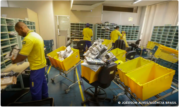 Funcionários dos Correios organizam encomendas em carrinhos amarelos dentro de um centro de triagem.