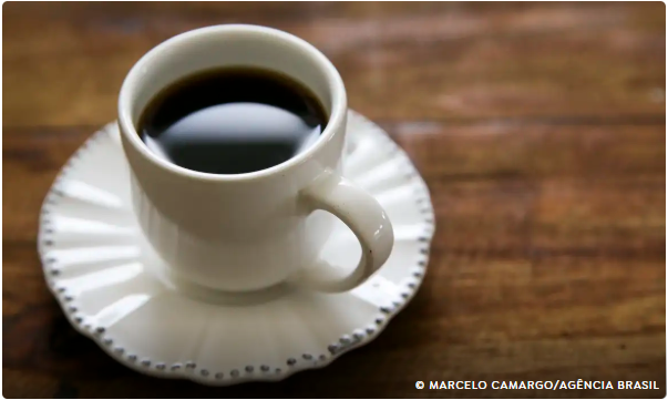 Xícara de café preto sobre pires branco, apoiada em mesa de madeira.