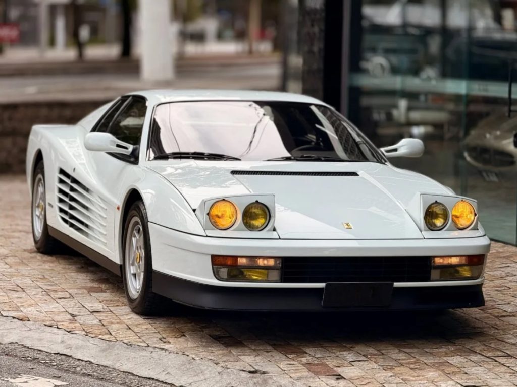 Ferrari Testarossa 1988 branca estacionada em área externa, vista de frente, com faróis escamoteáveis abertos e lentes amarelas.