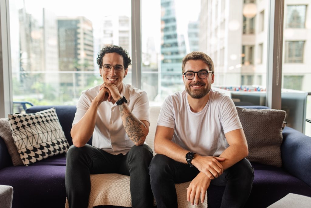 Dois profissionais sentados em sofá de escritório moderno, sorrindo, com vista urbana ao fundo.