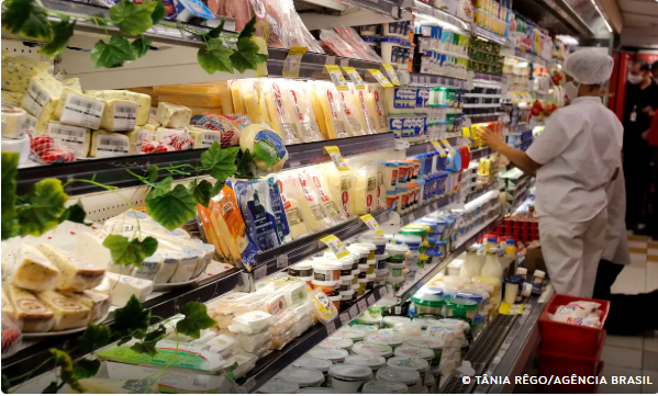 Funcionário organiza produtos refrigerados em prateleiras de supermercado, que exibem laticínios e itens de mercearia.
