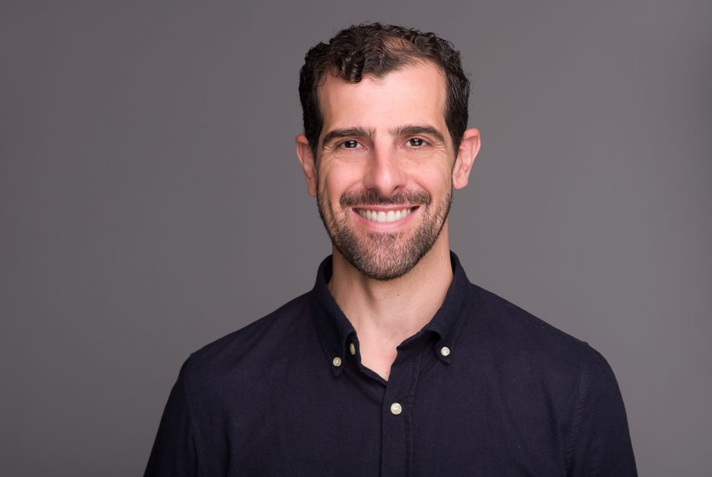 Retrato profissional de homem sorrindo em estúdio, usando camisa azul-marinho, sobre fundo cinza.